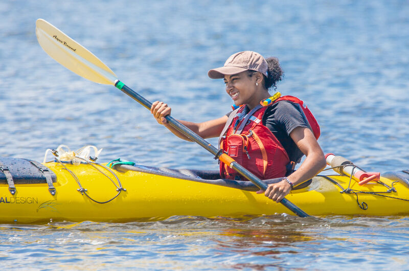 The image shows a young woman kayaking on a sunny day. She is wearing a life vest and a cap, holding a paddle, and smiling. The kayak is yellow, and the water is calm. The overall scene suggests a pleasant outdoor activity.